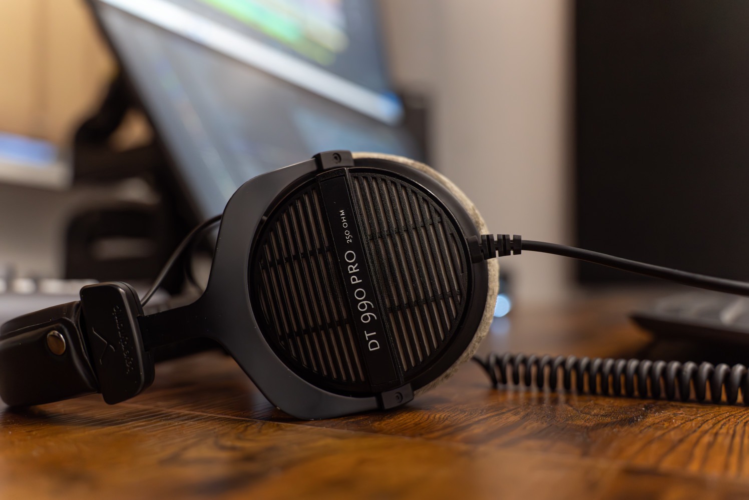 Profesjonalne słuchawki studyjne DT 990 PRO 250 Ohm na drewnianym biurku, z widocznym kablem spiralnym i fragmentem monitora w tle. Sprzęt audio dla studia.