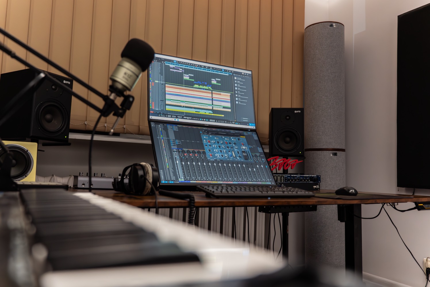 Profesjonalne stanowisko do miksowania dźwięku z dwoma monitorami, mikrofonem pojemnościowym i klawiaturą MIDI. Widoczne głośniki studyjne i panele akustyczne.