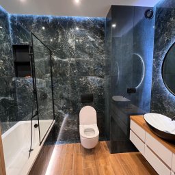 AMAT Usługi Remontowo-Budowlane - Nowoczesna łazienka z czarnymi płytkami imitującymi marmur, drewnopodobną podłogą i podświetleniem LED. Wanna z czarną kabiną, umywalka nablatowa i okrągłe lustro.