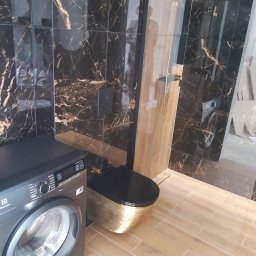 AMAT Usługi Remontowo-Budowlane - Elegancka łazienka z czarno-złotymi płytkami imitującymi marmur, złotą armaturą i toaletą. Widoczna pralka Electrolux. Podłoga wyłożona płytkami imitującymi drewno.