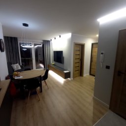 AMAT Usługi Remontowo-Budowlane - Nowoczesne wykończenie wnętrza salonu w Starachowicach: drewniana podłoga, oświetlenie LED, minimalistyczny design, dębowe drzwi i telewizor na ścianie. Przytulna aranżacja.