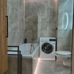 AMAT Usługi Remontowo-Budowlane - Nowoczesna łazienka w szarościach z podświetlaną wanną, pralką i toaletą. Minimalistyczny design, płytki imitujące beton, oświetlenie LED.