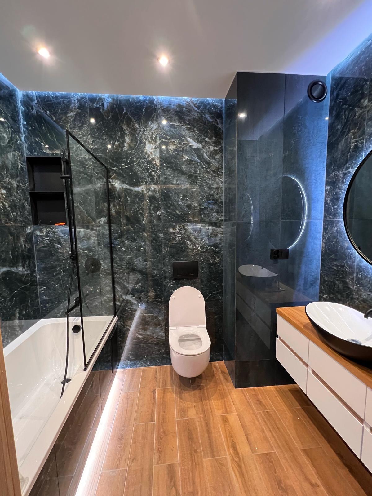 Nowoczesna łazienka z czarnymi płytkami imitującymi marmur, drewnopodobną podłogą i podświetleniem LED. Wanna z czarną kabiną, umywalka nablatowa i okrągłe lustro.