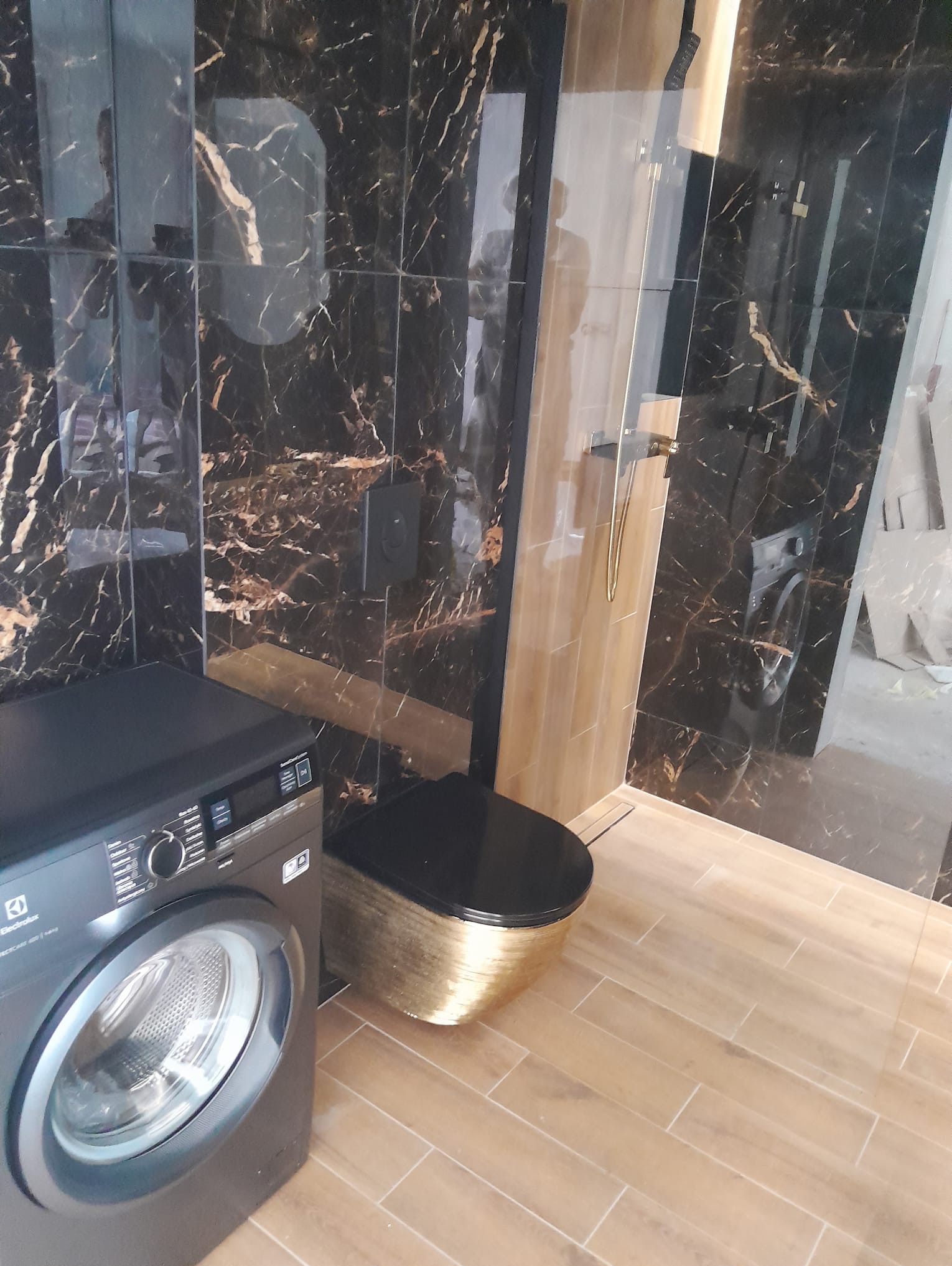 Elegancka łazienka z czarno-złotymi płytkami imitującymi marmur, złotą armaturą i toaletą. Widoczna pralka Electrolux. Podłoga wyłożona płytkami imitującymi drewno.