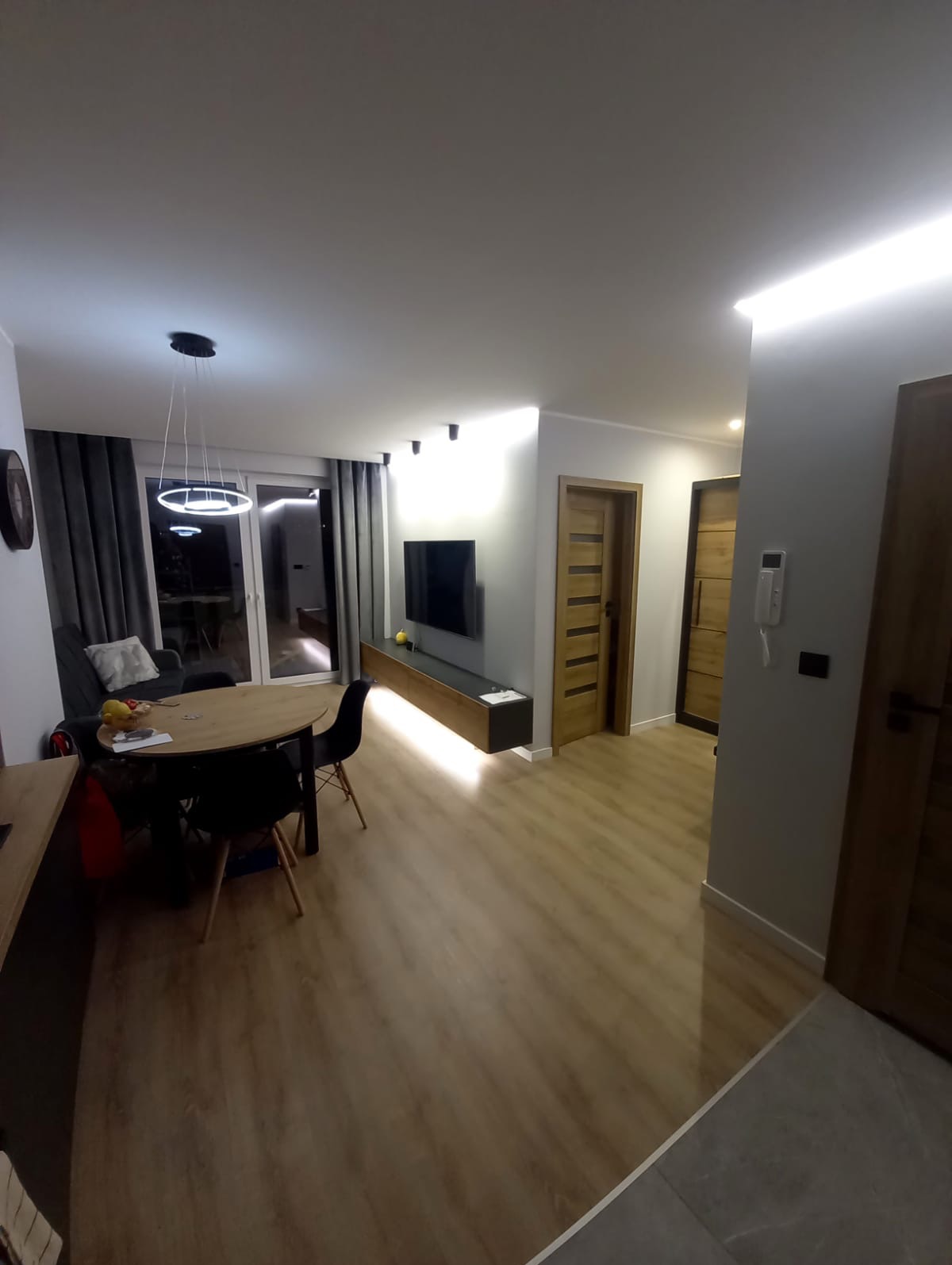 Nowoczesne wykończenie wnętrza salonu w Starachowicach: drewniana podłoga, oświetlenie LED, minimalistyczny design, dębowe drzwi i telewizor na ścianie. Przytulna aranżacja.