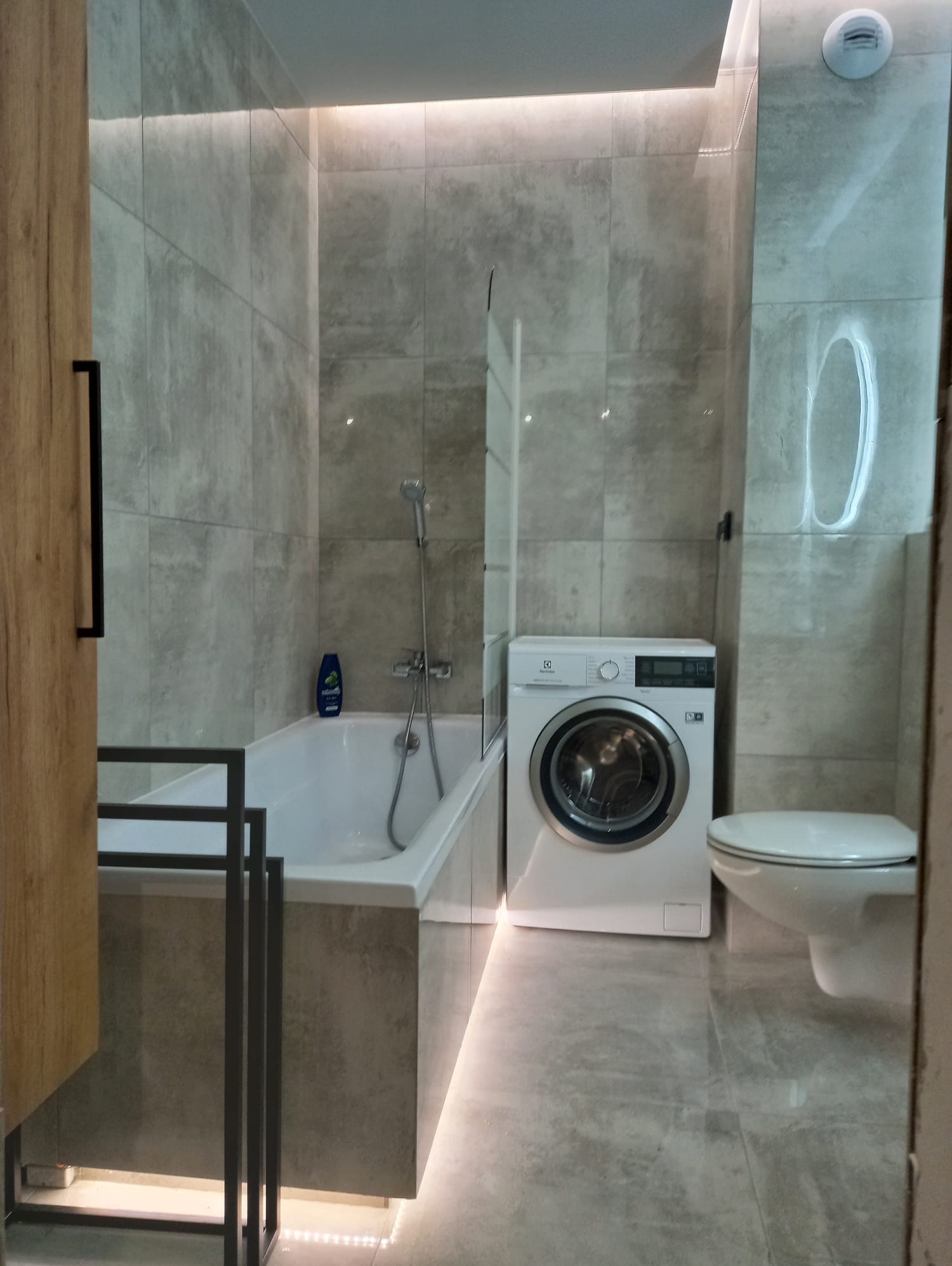 Nowoczesna łazienka w szarościach z podświetlaną wanną, pralką i toaletą. Minimalistyczny design, płytki imitujące beton, oświetlenie LED.