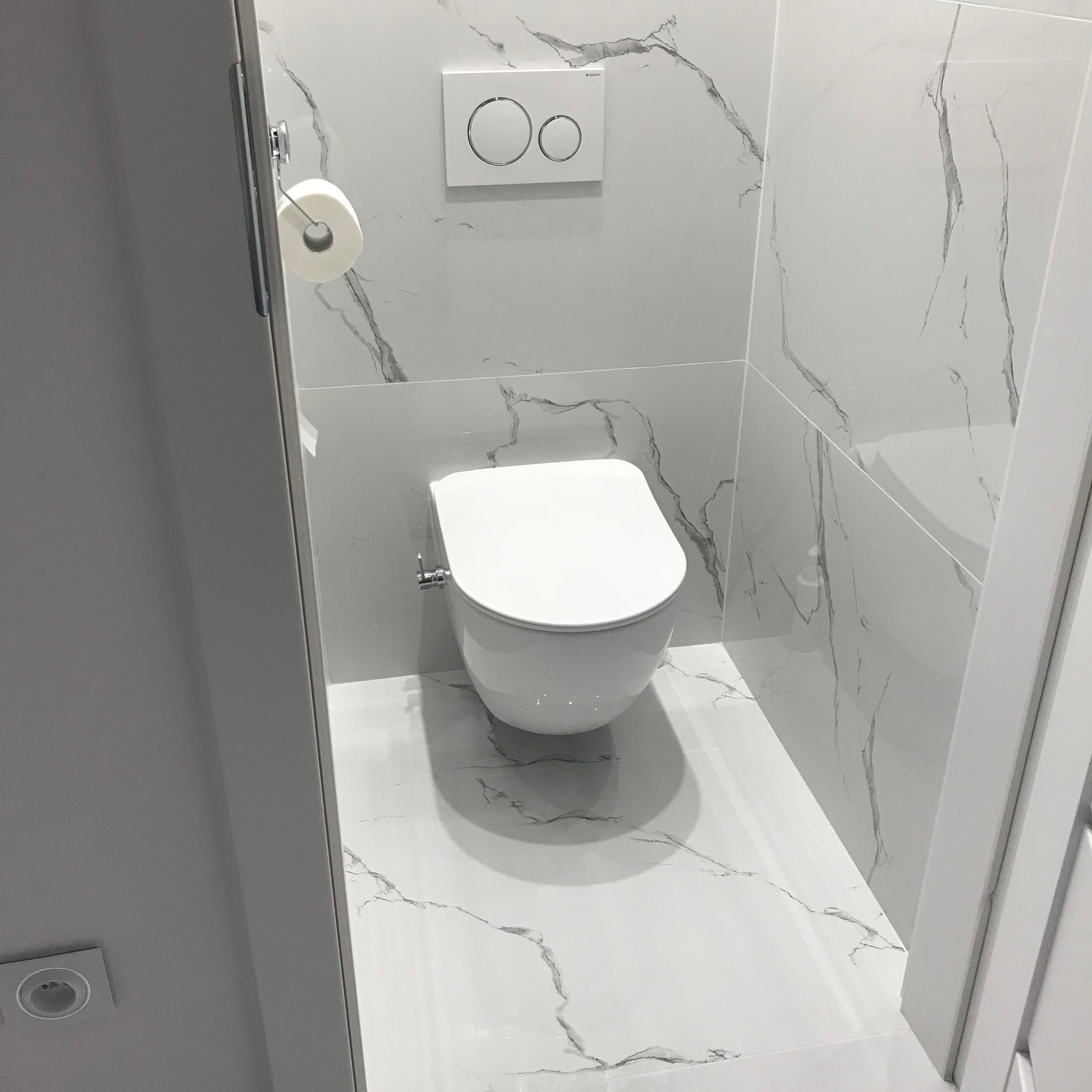 Biała toaleta wisząca w nowoczesnej łazience wykończonej płytkami imitującymi marmur. Widoczny uchwyt na papier toaletowy i przycisk spłuczki.