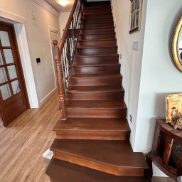 FRWD SPÓŁKA Z OGRANICZONĄ ODPOWIEDZIALNOŚCIĄ - Wysokie, drewniane schody z czarną balustradą w jasnym wnętrzu domu. Widok od dołu, ukazujący stopnie i fragment parkietu. Dekoracje na ścianie.