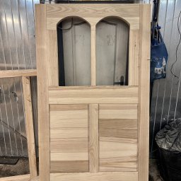 FRWD Craft Manufacturing - Surowe, drewniane drzwi wewnętrzne z szybą w stylu retro, w warsztacie w Krakowie. Widoczna struktura drewna i niedokończone wykończenie. Obok leży rama drzwi.