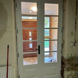 FRWD SPÓŁKA Z OGRANICZONĄ ODPOWIEDZIALNOŚCIĄ - Białe, przeszklone drzwi dwuskrzydłowe w trakcie montażu, widoczne surowe ściany i instalacje. W tle widoczne kolejne drzwi i fragment ogrodu.