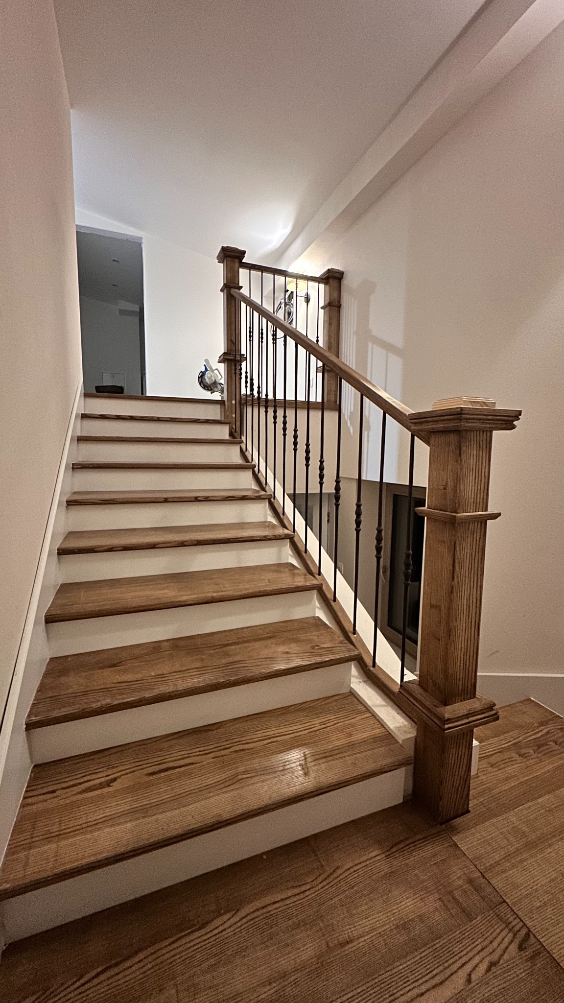 Drewniane schody z białymi podstopnicami i czarnymi balustradami, oświetlone ciepłym światłem. Widoczne detale wykończenia i naturalna tekstura drewna.