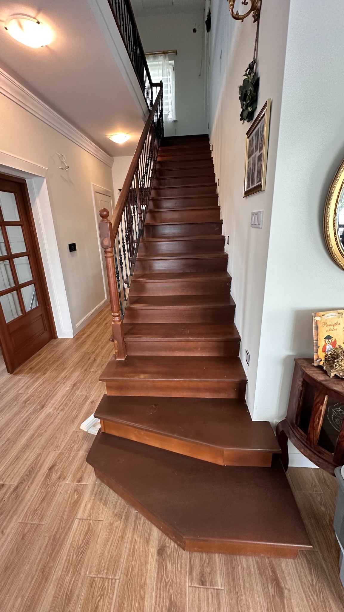 Wysokie, drewniane schody z czarną balustradą w jasnym wnętrzu domu. Widok od dołu, ukazujący stopnie i fragment parkietu. Dekoracje na ścianie.