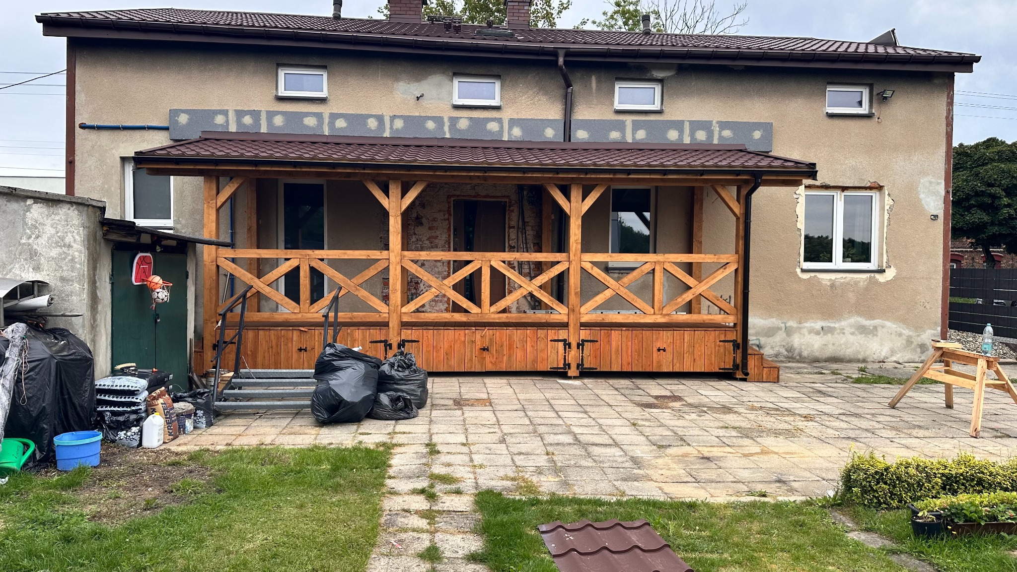 Drewniana pergola z balustradą w rustykalnym stylu, przylegająca do budynku z widocznymi śladami prac remontowych. Uwagę zwracają czarne worki na śmieci przed konstrukcją.