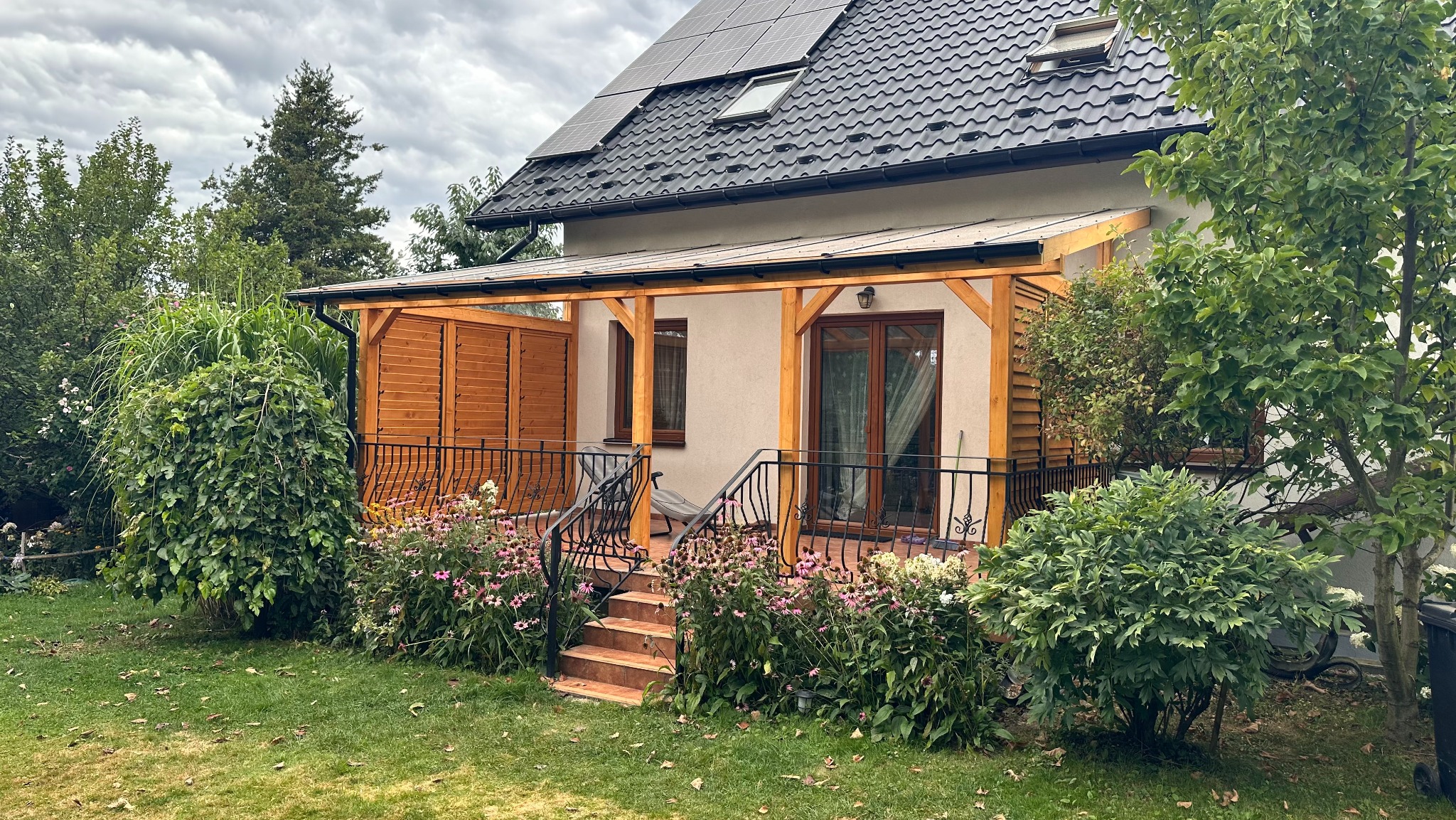 Zadaszony taras drewniany z balustradą, schodami i roślinnością. Widoczne panele słoneczne na dachu domu. Ujęcie z poziomu ogrodu, pochmurne niebo w tle.