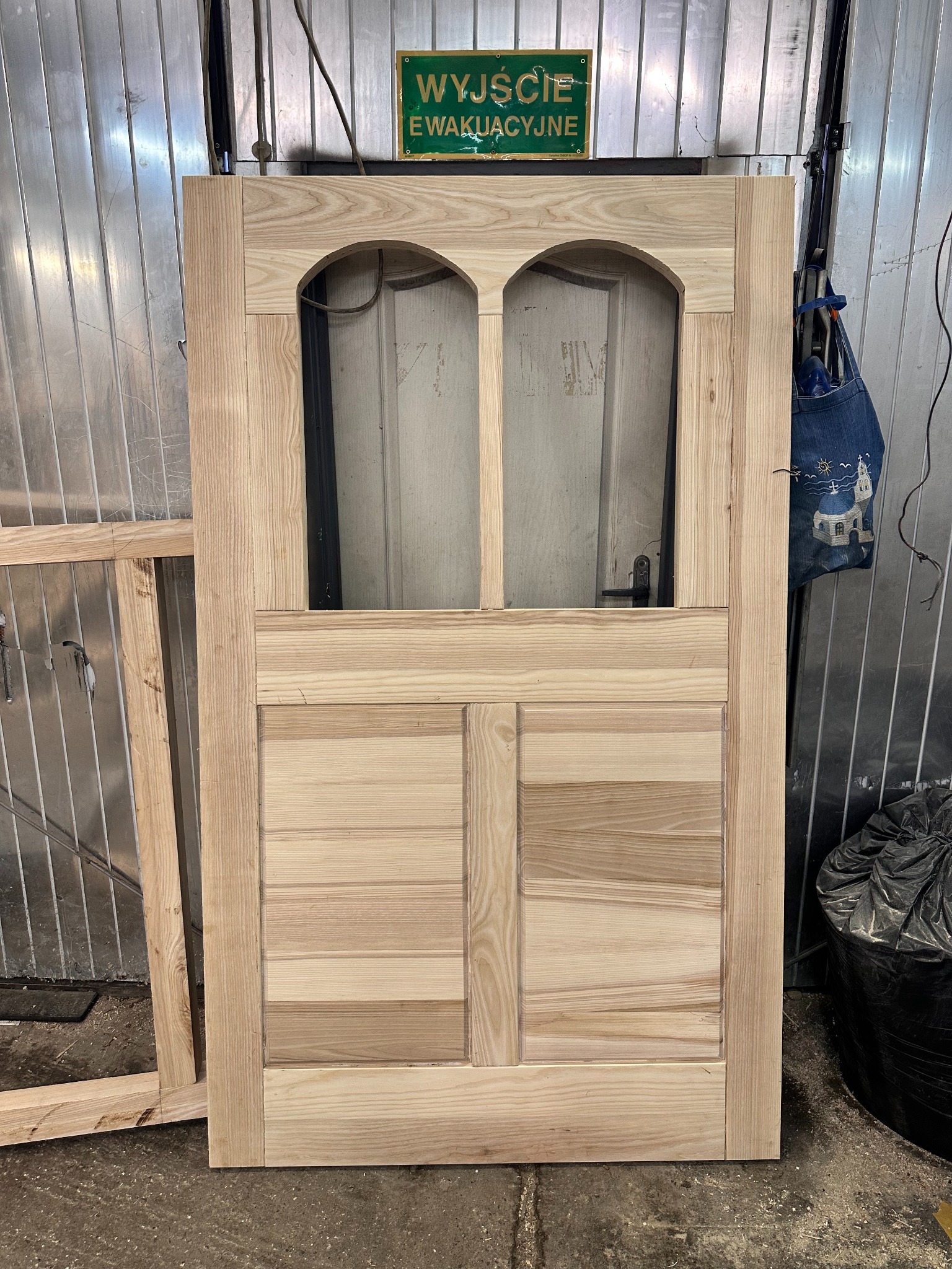 Surowe, drewniane drzwi wewnętrzne z szybą w stylu retro, w warsztacie w Krakowie. Widoczna struktura drewna i niedokończone wykończenie. Obok leży rama drzwi.