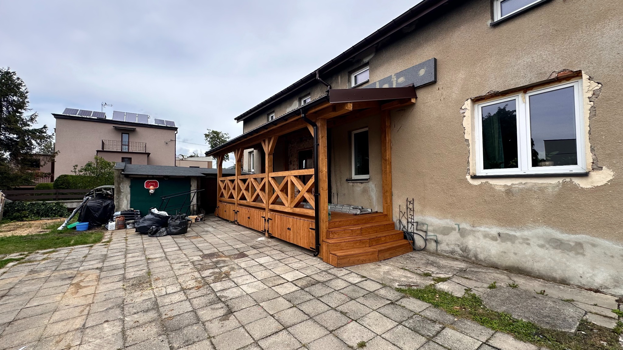 Drewniana zabudowa tarasu z balustradą krzyżową i schodami, widoczna z zewnątrz budynku. Nowe okno w elewacji. Na podwórku worki i narzędzia.