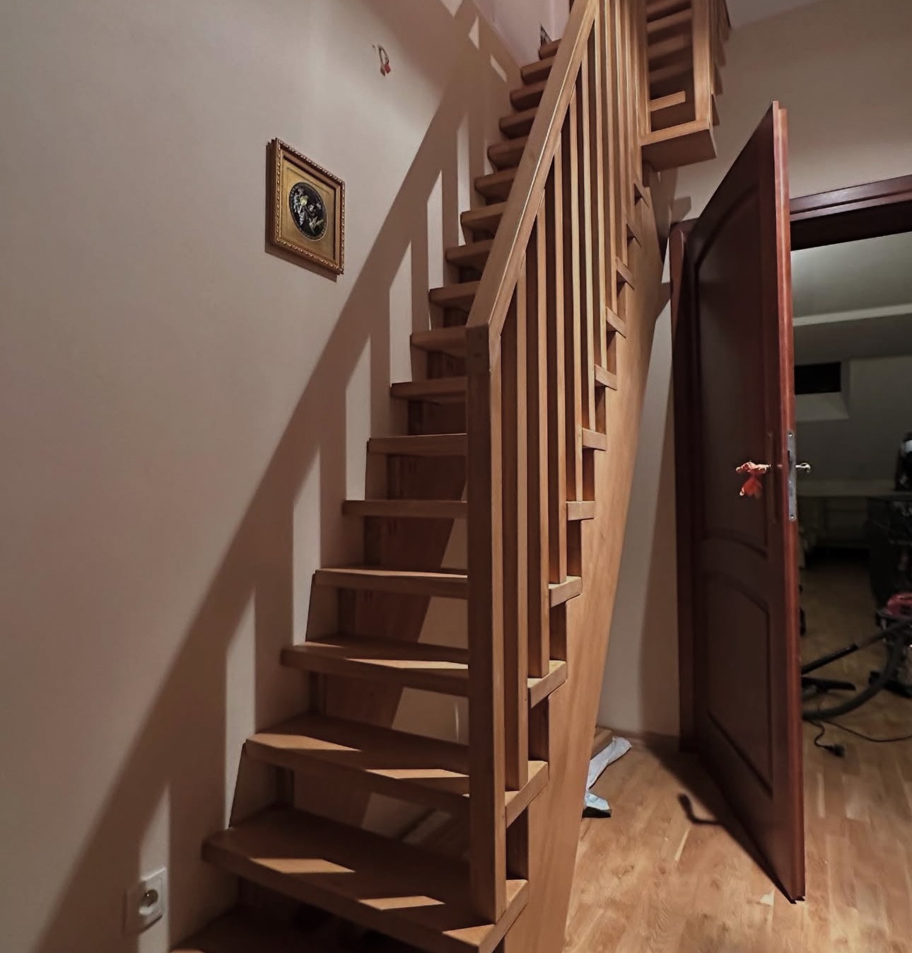 Drewniane schody z balustradą, prowadzące na górę, z widoczną otwartą, ciemną futryną drzwiową i obrazkiem na ścianie. Naturalne oświetlenie podkreśla strukturę drewna.