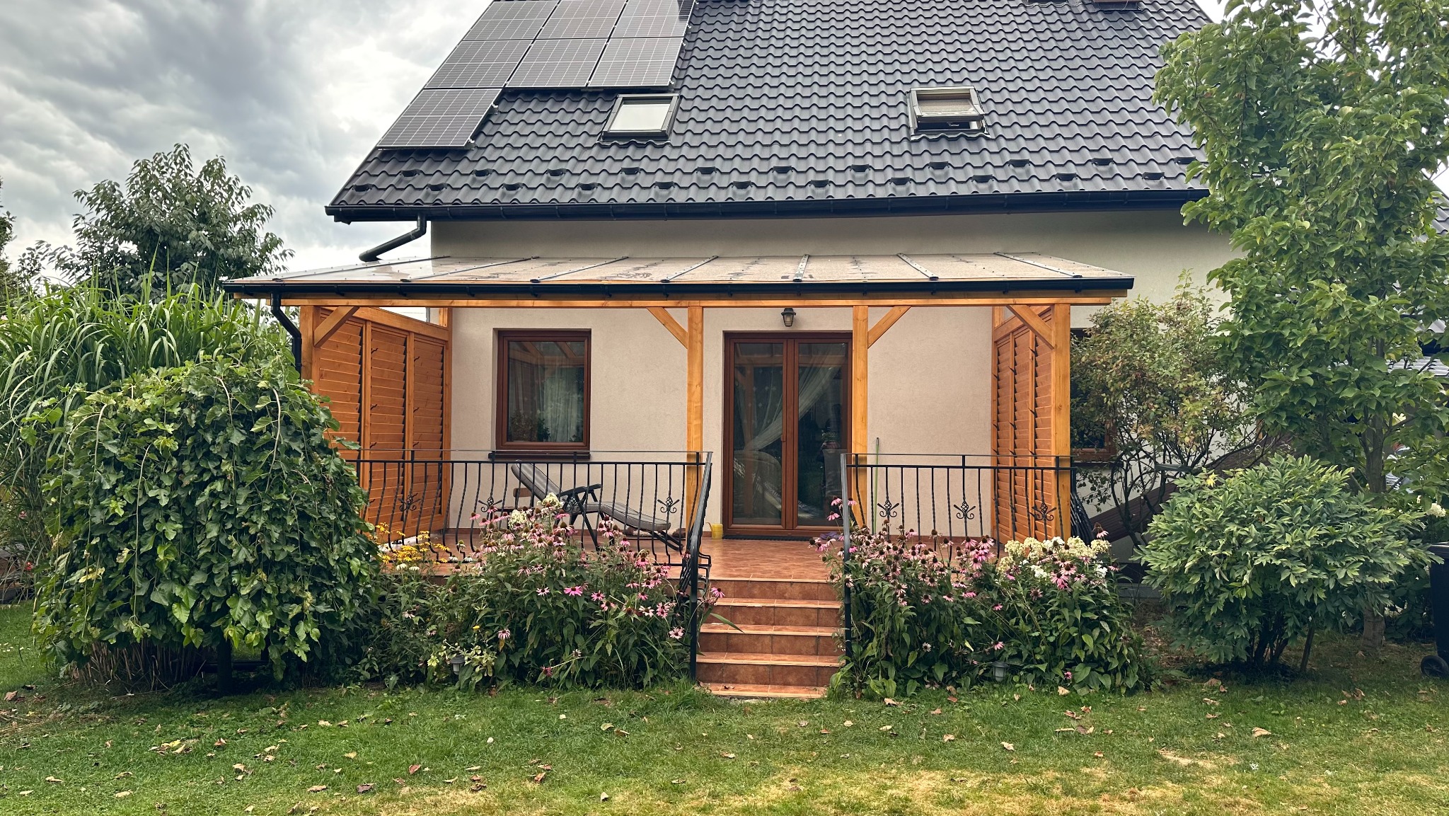 Zadaszony taras z drewnianą konstrukcją i schodami, otoczony zielenią. Widoczne panele słoneczne na dachu domu. Uroku dodają kwiaty wzdłuż balustrady.