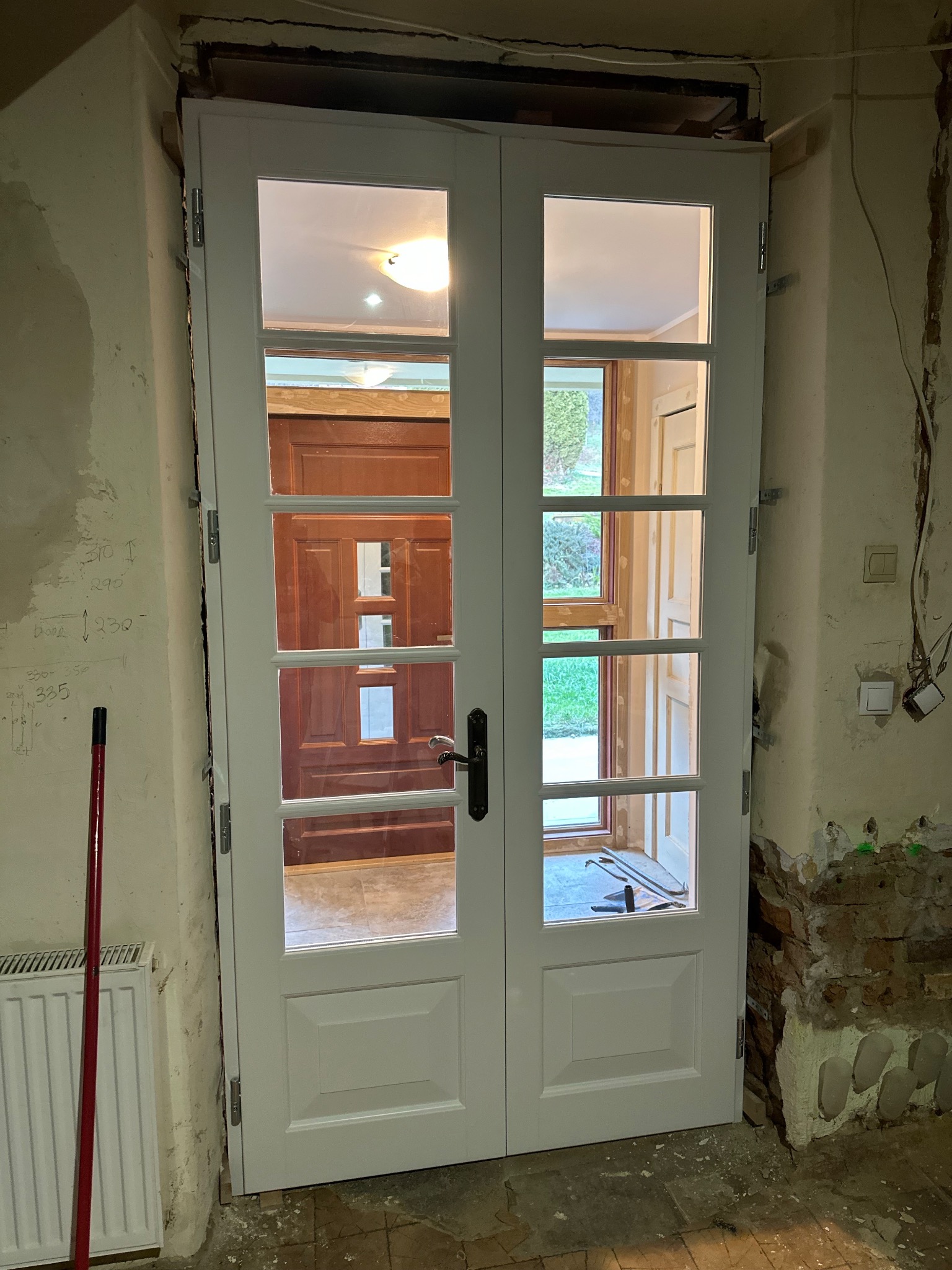 Białe, przeszklone drzwi dwuskrzydłowe w trakcie montażu, widoczne surowe ściany i instalacje. W tle widoczne kolejne drzwi i fragment ogrodu.