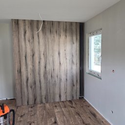 Design flow - Montaż drewnianej szafy wnękowej z panelami imitującymi drewno, widoczne panele podłogowe i odkurzacz przemysłowy. Szare ściany, okno z parapetem.