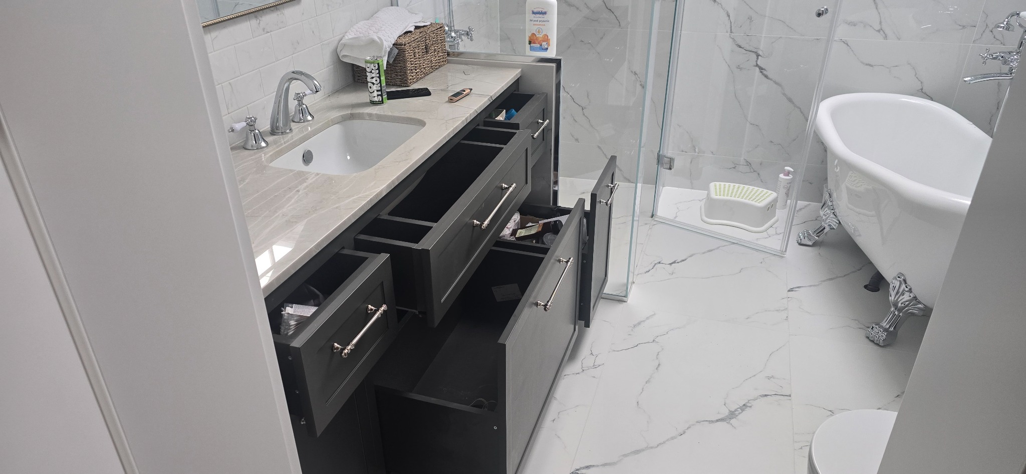 Nowoczesna łazienka z szarą szafką pod umywalką z otwartymi szufladami, marmurowym blatem i białą wanną na ozdobnych nóżkach. Widoczne detale armatury i akcesoriów.