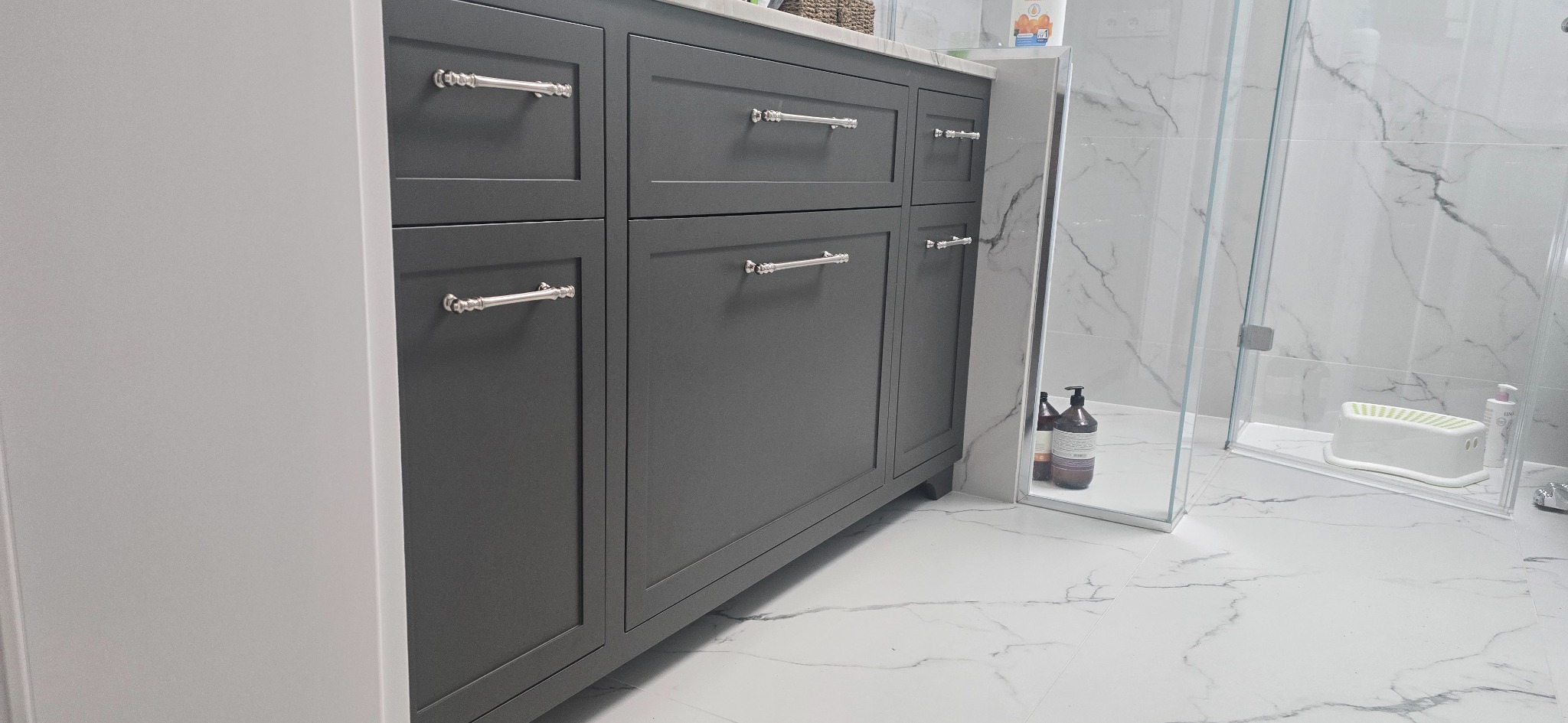 Szara szafka łazienkowa z chromowanymi uchwytami na tle marmurowych płytek i kabiny prysznicowej. Minimalistyczny design, nowoczesne wykończenie.