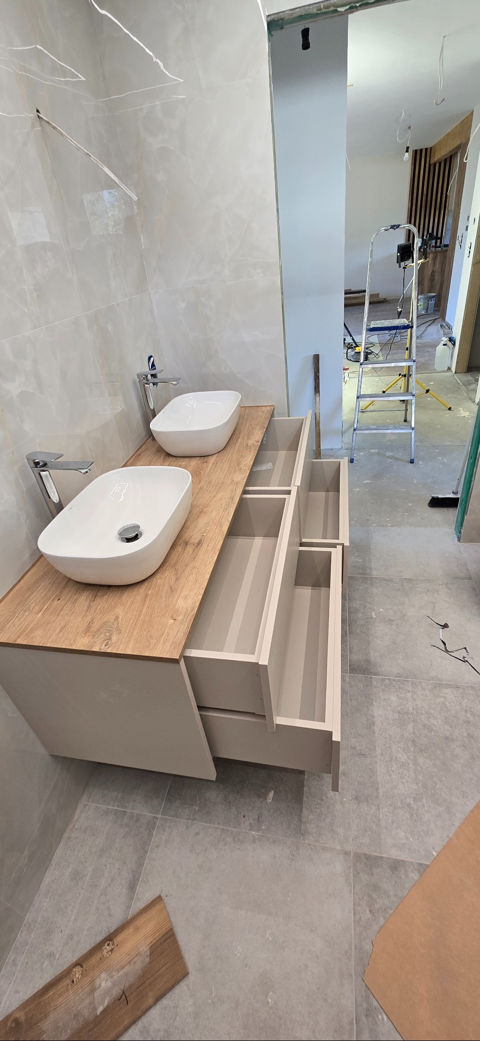 Nowoczesna szafka łazienkowa z drewnianym blatem i dwiema umywalkami. Otwarta szuflada ukazuje wnętrze. W tle drabina i elementy wykończenia wnętrza.