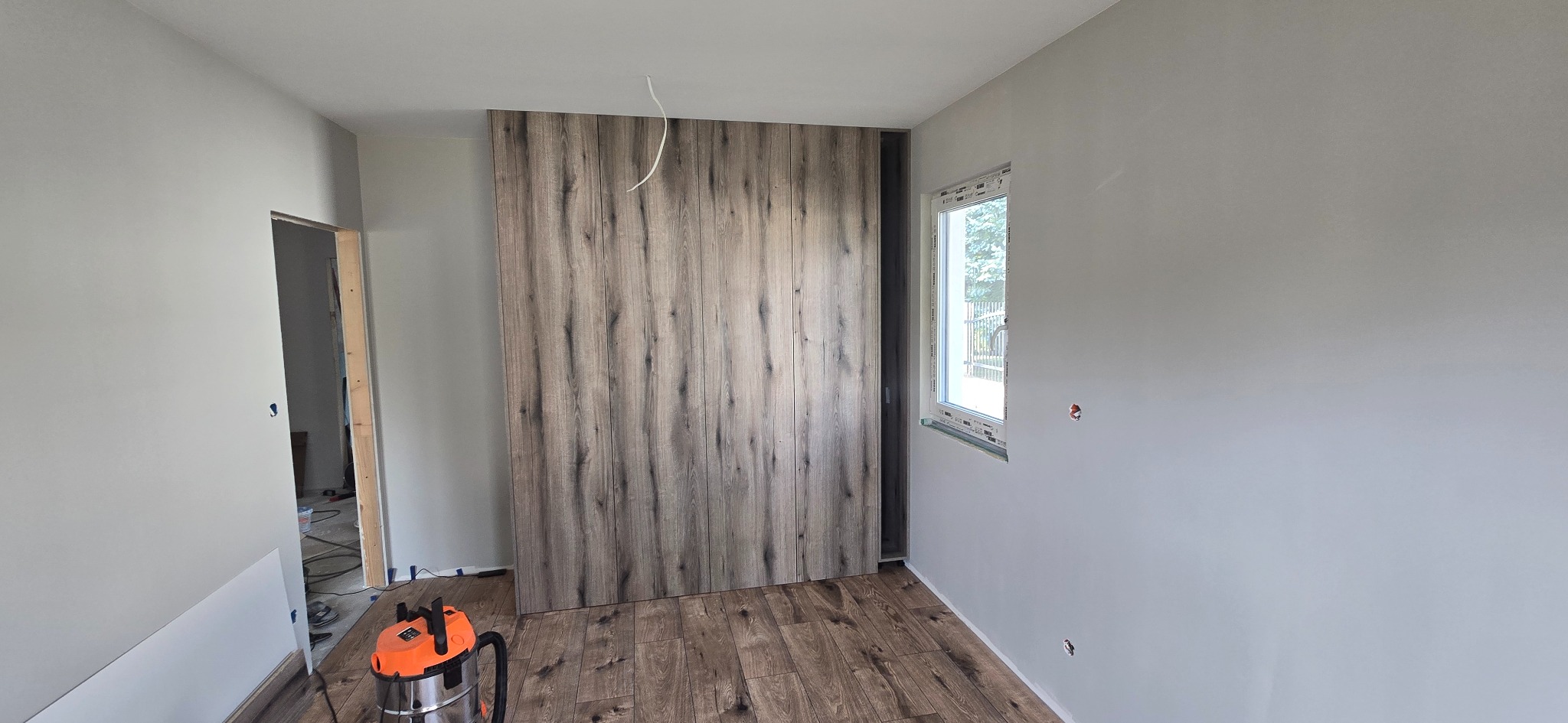 Montaż drewnianej szafy wnękowej z panelami imitującymi drewno, widoczne panele podłogowe i odkurzacz przemysłowy. Szare ściany, okno z parapetem.