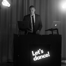 TOMCZAK FOTO BARTOSZ TOMCZAK - Czarno-białe zdjęcie DJ-a w garniturze za konsoletą z napisem 'Let's dance!', oświetlonego reflektorami, z dwoma laptopami. Profesjonalna oprawa muzyczna imprezy.