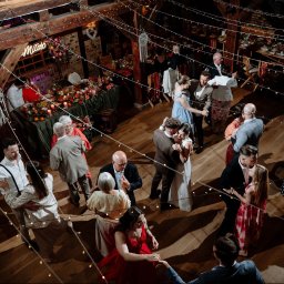 TOMCZAK FOTO BARTOSZ TOMCZAK - Parkiet w rustykalnej sali weselnej wypełniony tańczącymi parami, dekoracje kwiatowe i girlandy świetlne tworzą ciepłą atmosferę. Ujęcie z góry.