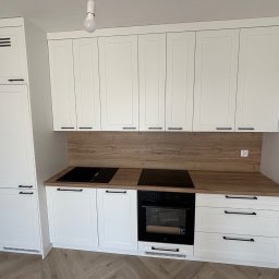 DNS Meb - Biała zabudowa kuchenna z drewnianym blatem i panelem, czarne uchwyty i płyta indukcyjna. Nowoczesny design, jasne wnętrze, parkiet w jodełkę.