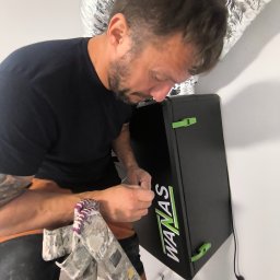 Reku Masters Sp. z o.o. - Technik na drabinie, z tatuażami, instaluje lub serwisuje czarne urządzenie WANAS z zielonymi zatrzaskami, podłączone do srebrnego kanału wentylacyjnego.