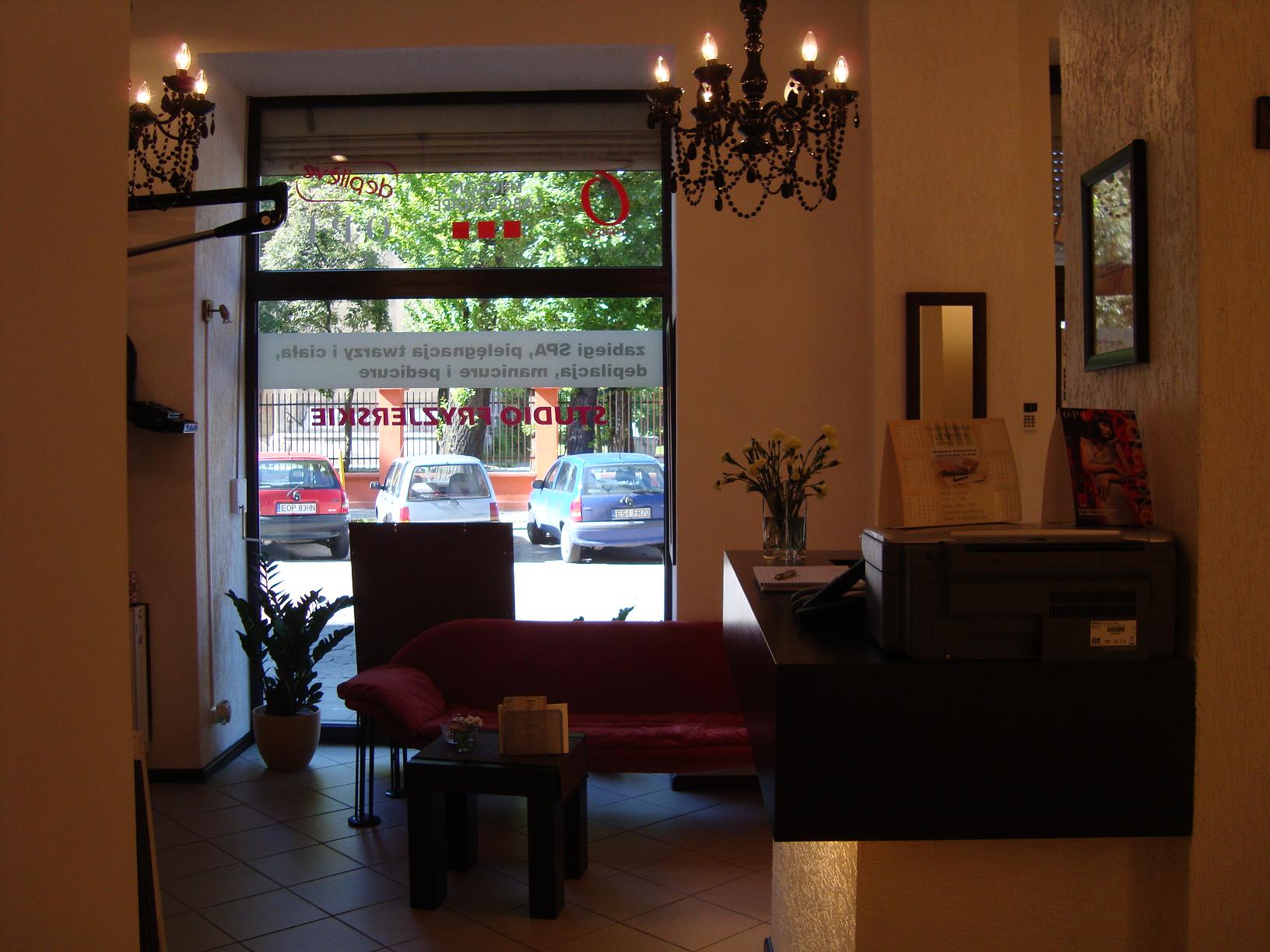 Wnętrze poczekalni: czerwona sofa, stolik kawowy z kwiatami, recepcja z drukarką, żyrandol w stylu retro, widok na ulicę przez przeszklone drzwi z napisem 'Studio Kosmetyczne'.