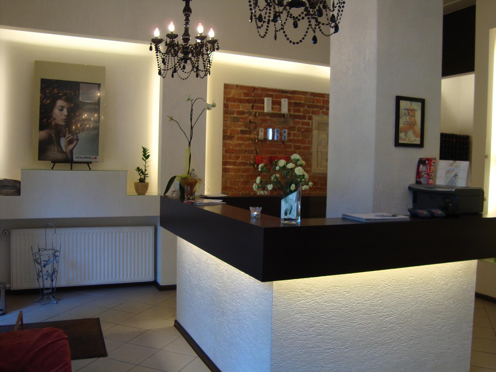 Recepcja w salonie, z ciemnym blatem i podświetlanym, teksturowanym frontem, dekoracje kwiatowe, żyrandole i plakat Goldwell w tle.