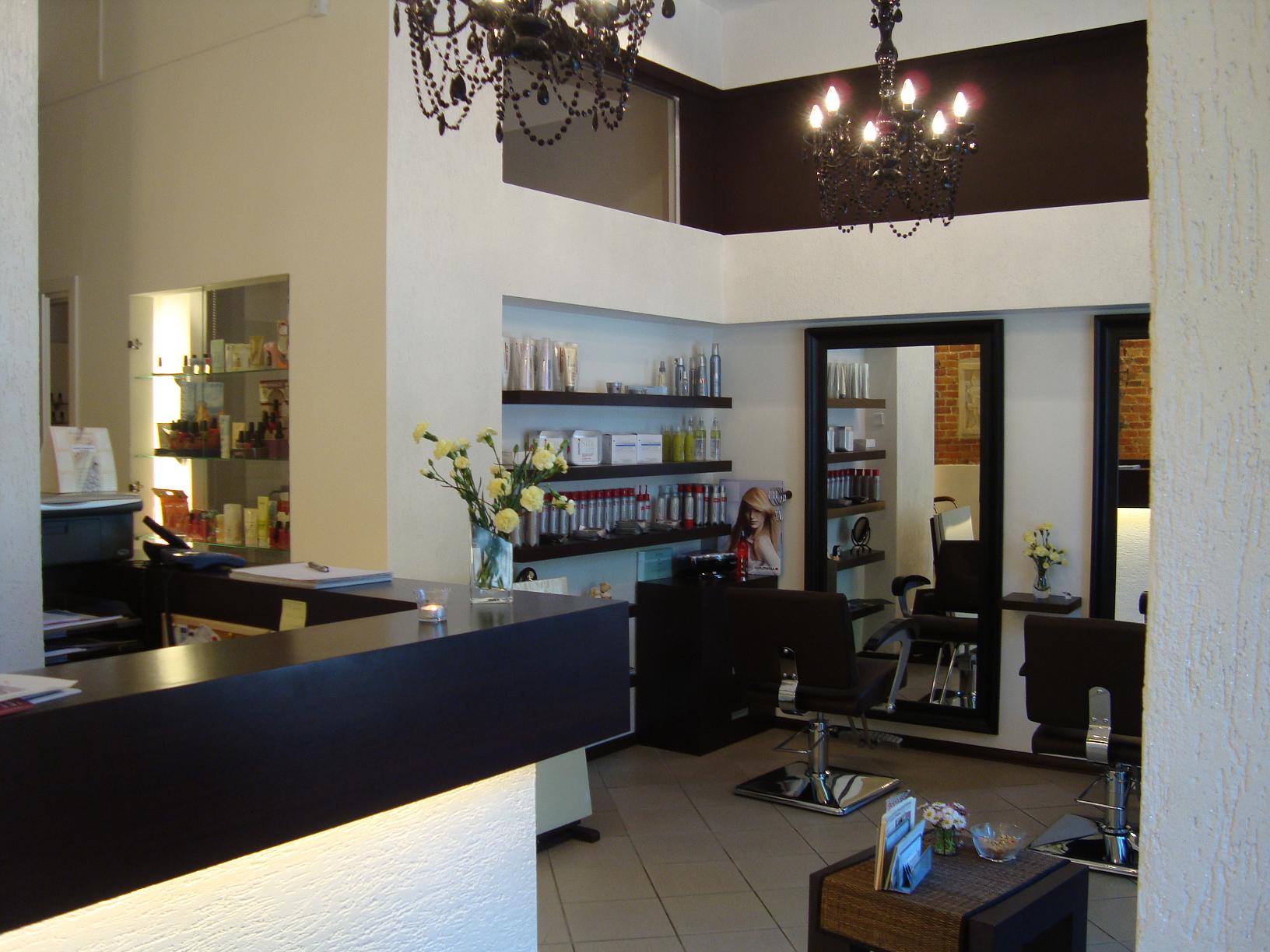 Wnętrze salonu fryzjerskiego z recepcją, ciemnymi meblami, lustrami i dekoracyjnymi żyrandolami, na półkach widoczne kosmetyki i produkty do stylizacji włosów.