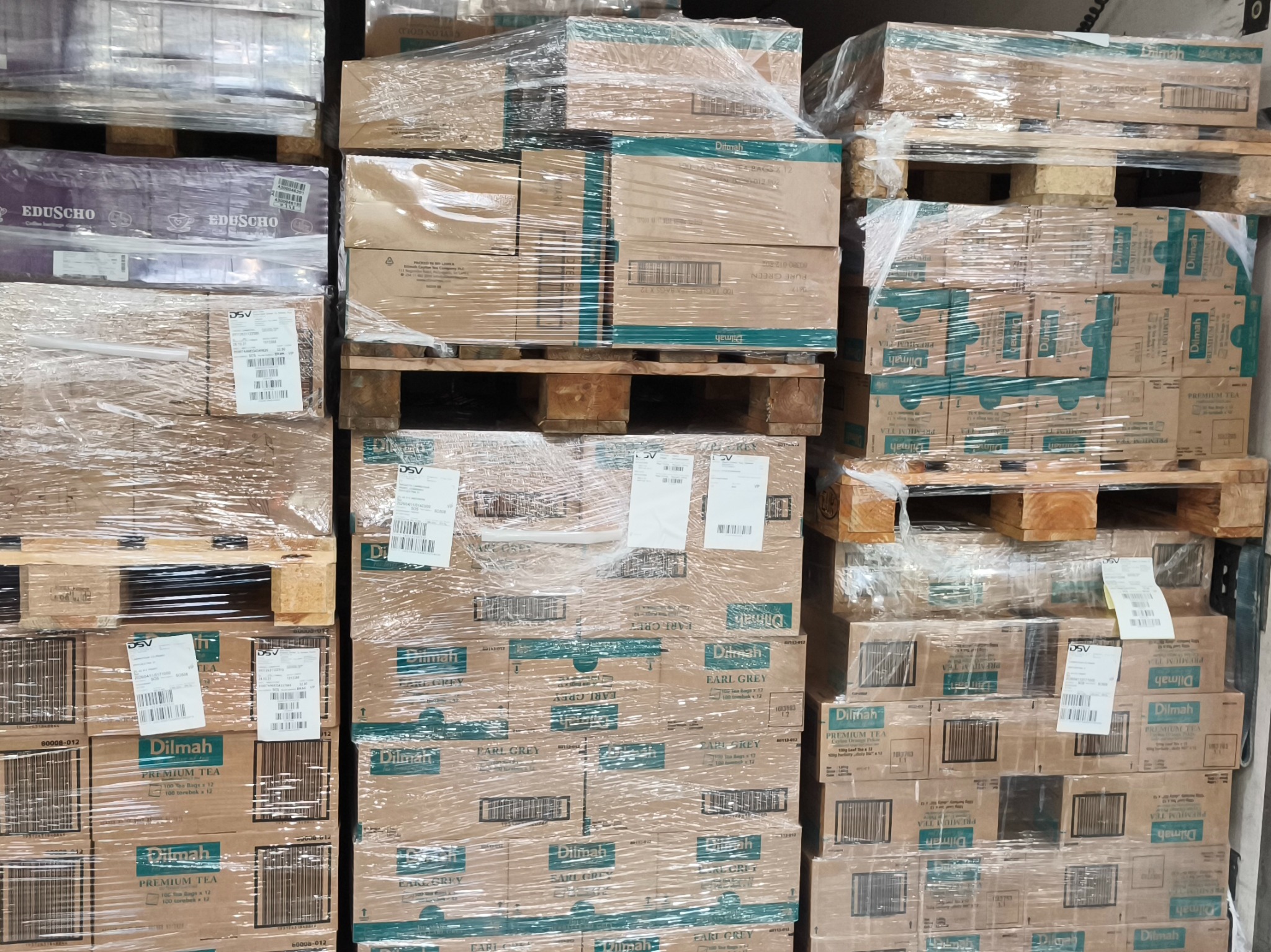 Palety z kartonami herbaty Dilmah i kawy Eduscho, owinięte folią stretch, gotowe do transportu. Widoczne etykiety DSV. Magazyn z towarem.