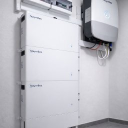 AIR SOUL SPÓŁKA Z OGRANICZONĄ ODPOWIEDZIALNOŚCIĄ - Biały, modułowy magazyn energii Hoymiles zamontowany na ścianie, z widoczną skrzynką elektryczną i inwerterem. Minimalistyczne wnętrze z szarą podłogą.