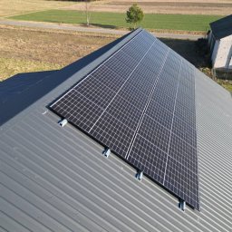 AIR SOUL SPÓŁKA Z OGRANICZONĄ ODPOWIEDZIALNOŚCIĄ - Panel słoneczny na szarym dachu z blachy trapezowej, widok z góry. W tle pole i budynek gospodarczy. Montaż paneli fotowoltaicznych na dachu.