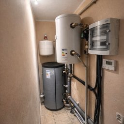 AIR SOUL SPÓŁKA Z OGRANICZONĄ ODPOWIEDZIALNOŚCIĄ - Zestaw zbiorników akumulacyjnych i rozdzielnica elektryczna w kotłowni. Widoczne rury instalacyjne i elementy sterujące. Ujęcie z dołu, podkreślające złożoność instalacji.