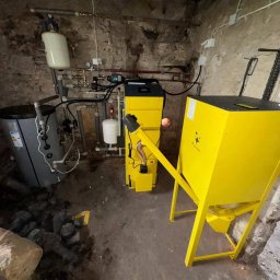 AIR SOUL SPÓŁKA Z OGRANICZONĄ ODPOWIEDZIALNOŚCIĄ - Żółty piec na paliwo stałe z podajnikiem ślimakowym w kotłowni o surowych ścianach. Widoczny zbiornik na wodę i instalacja. Ujęcie z bliska, z dołu.