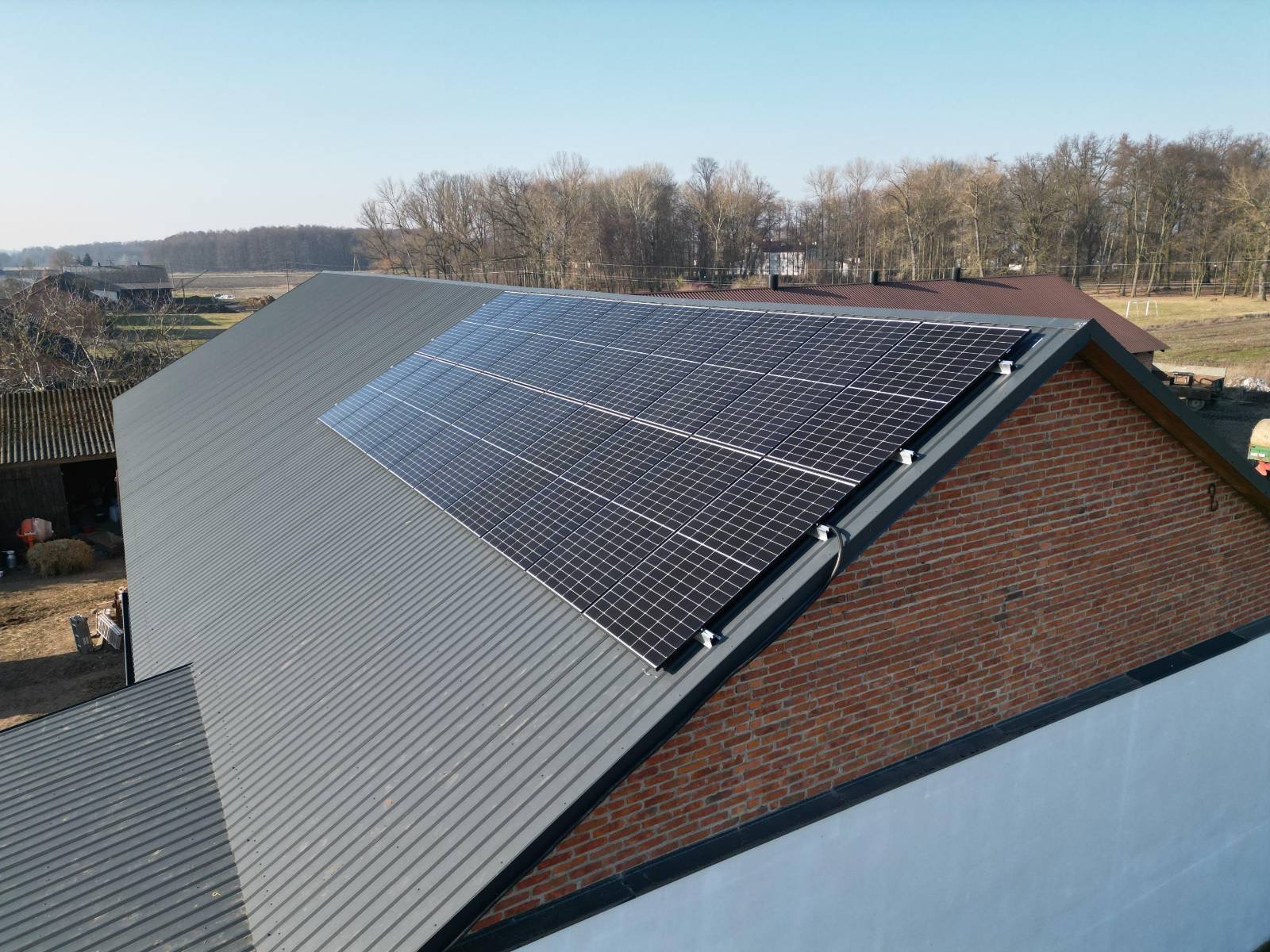 Panele słoneczne na szarym, blaszanym dachu budynku z ceglaną ścianą szczytową. Widok z góry, w tle drzewa i zabudowania wiejskie.