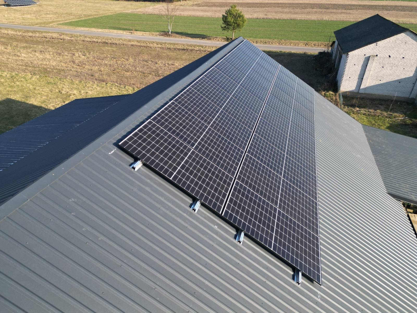Panel słoneczny na szarym dachu z blachy trapezowej, widok z góry. W tle pole i budynek gospodarczy. Montaż paneli fotowoltaicznych na dachu.