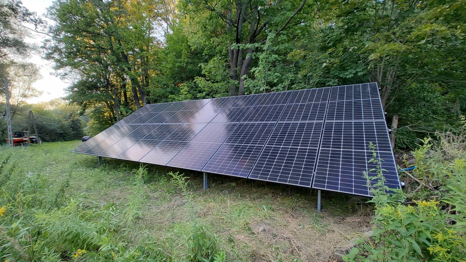 Instalacja fotowoltaiczna na gruncie, otoczona zielenią i drzewami. Panele ustawione pod kątem, widoczny traktor w tle. Ujęcie z bliska, naturalne oświetlenie.