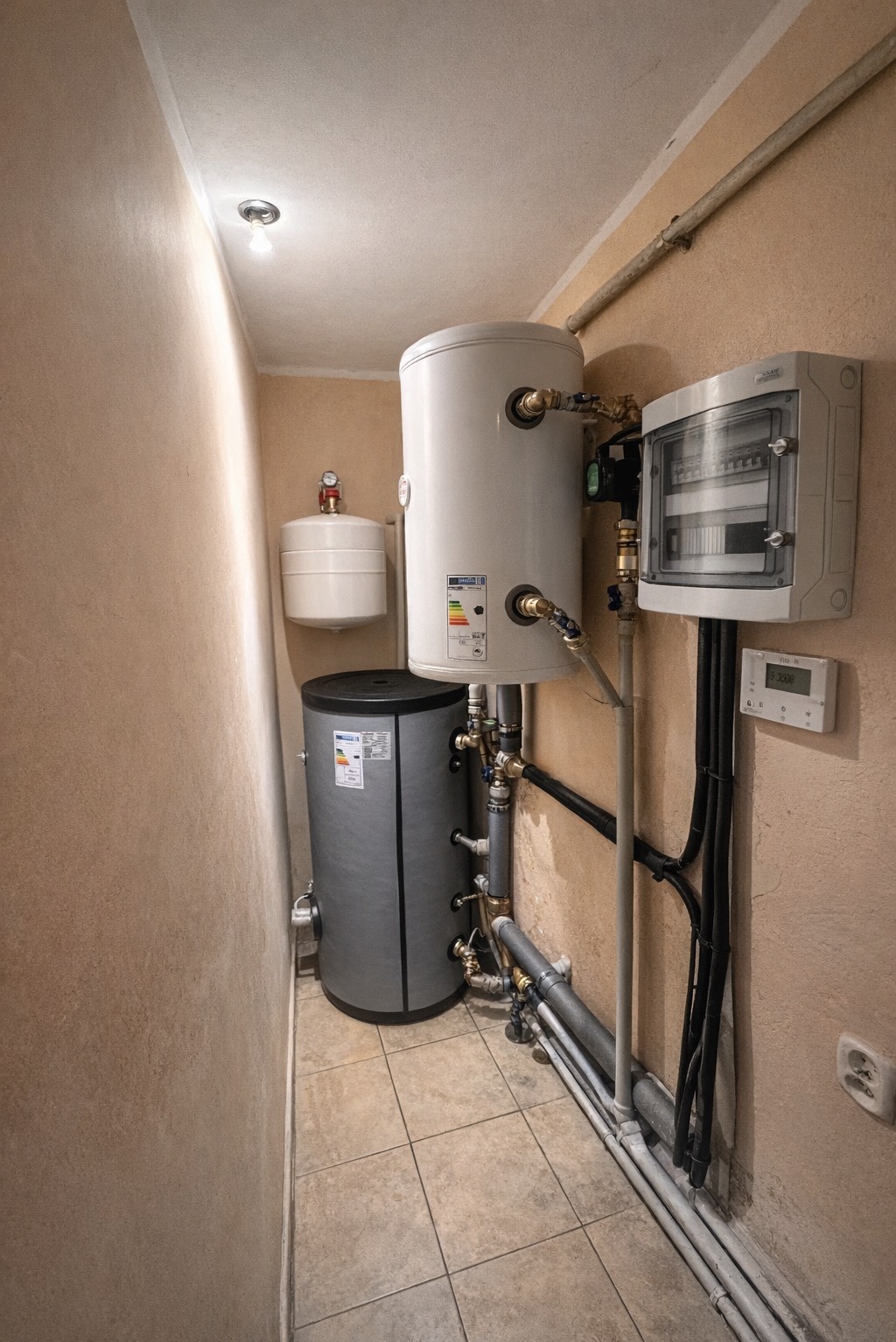 Zestaw zbiorników akumulacyjnych i rozdzielnica elektryczna w kotłowni. Widoczne rury instalacyjne i elementy sterujące. Ujęcie z dołu, podkreślające złożoność instalacji.