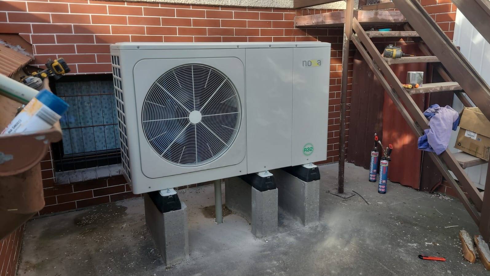 Zewnętrzna jednostka pompy ciepła Nova R32, zamontowana na betonowych blokach przy ceglanej ścianie. Widoczne narzędzia i schody w tle.