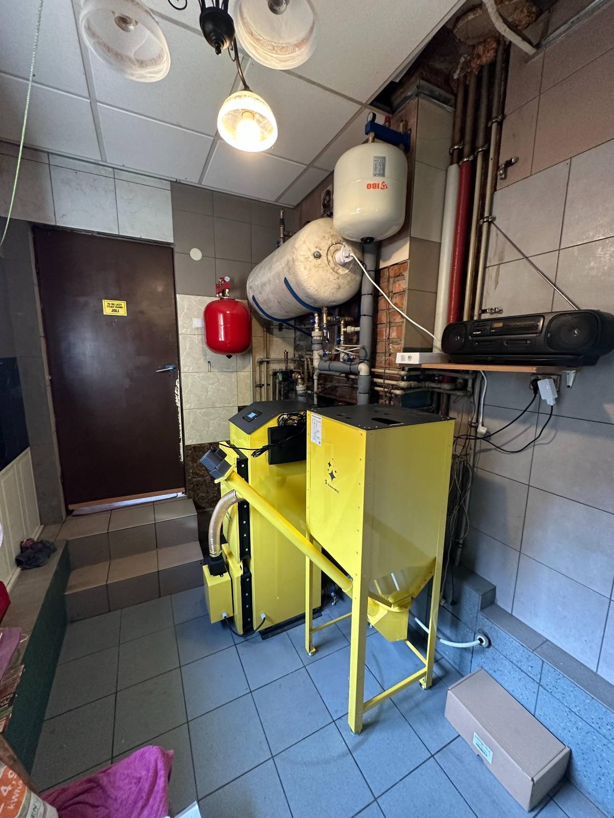 Żółty piec na paliwo stałe w kotłowni z szarymi płytkami, widoczne zbiorniki, rury i radio na półce. U góry lampa, z boku drzwi. Na podłodze karton.