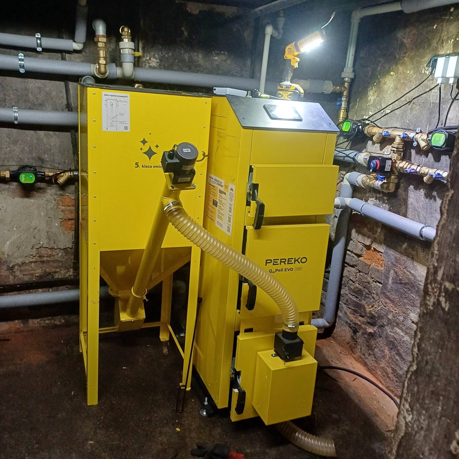 Żółty piec na pellet Pereko Q-Pell EVO 5. klasy emisji w kotłowni z widocznymi rurami instalacji grzewczej i oświetleniem roboczym.
