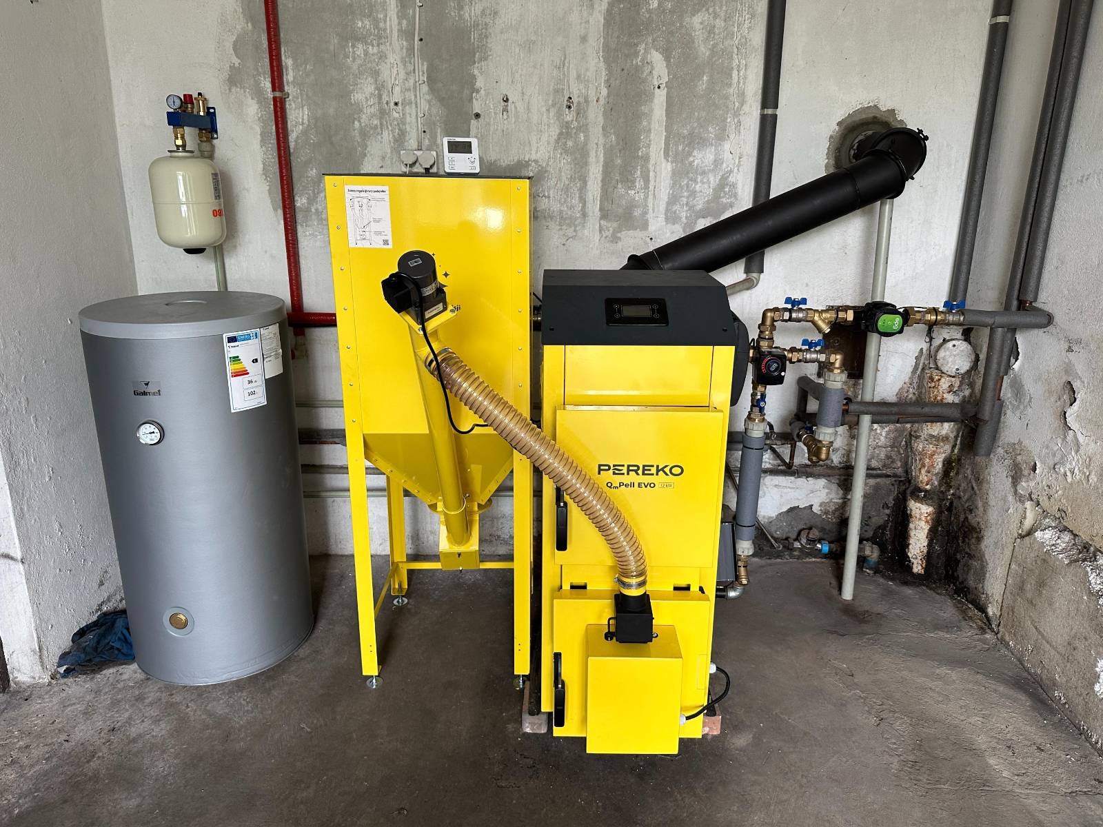 Żółty piec na pellet Pereko Q-Pell EVO z podajnikiem i zasobnikiem, szary bojler Galmet, instalacja CO w kotłowni z surowymi ścianami.