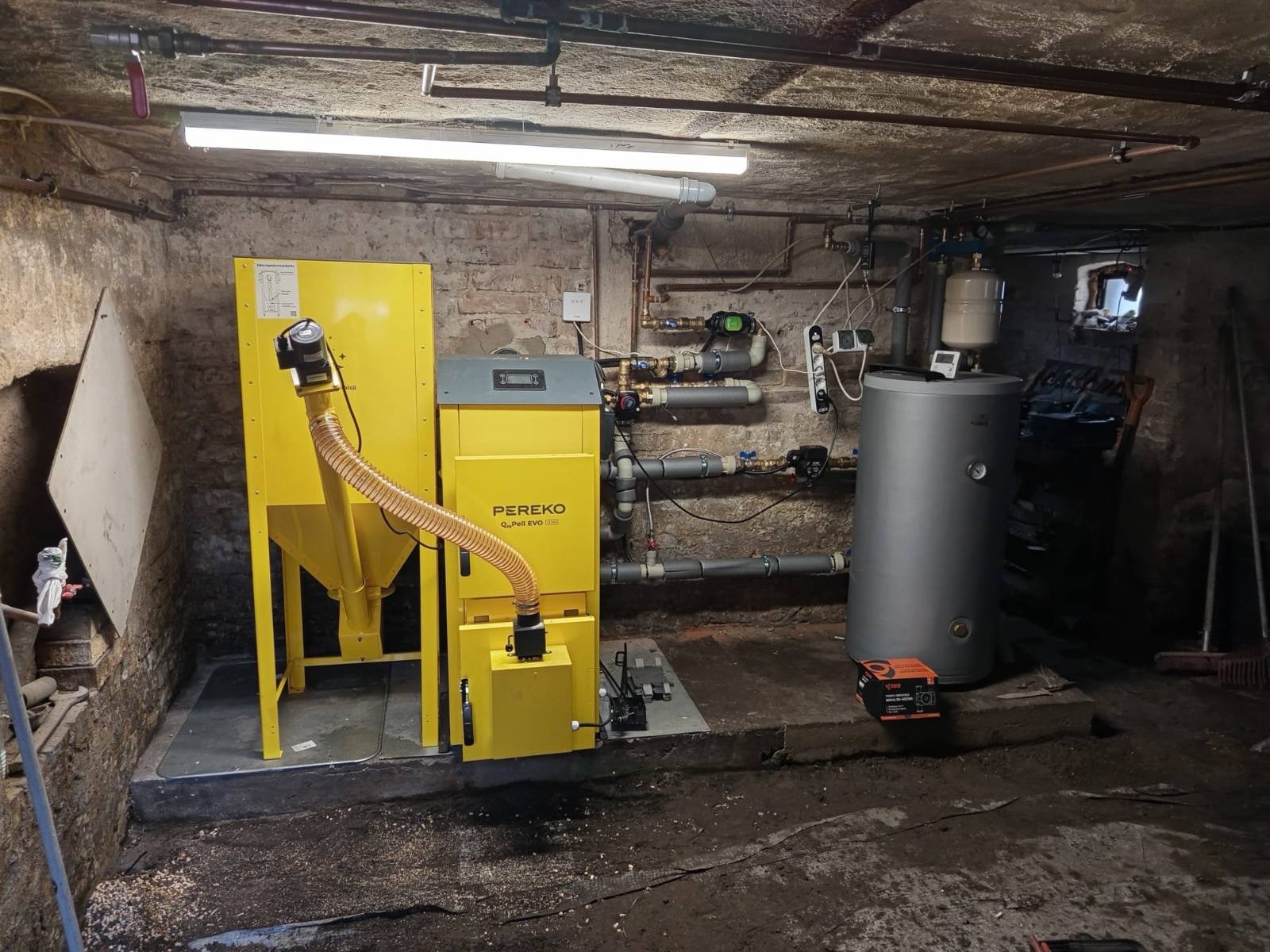 Żółty piec na pellet Pereko Q-Pell EVO 35 w piwnicy z instalacją grzewczą i zbiornikiem. Widoczne rury, zawory i osprzęt. Surowe ściany i podłoga.