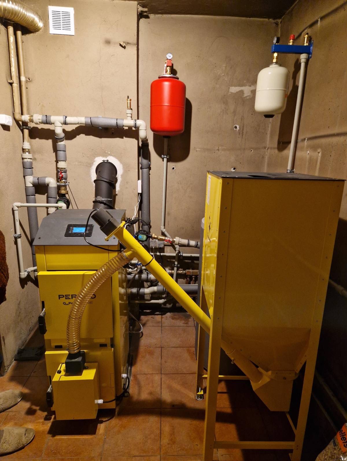 Żółty piec na pellet z podajnikiem w kotłowni, widoczne rury instalacji CO i zbiorniki wyrównawcze. Ujęcie z niskiego kąta, podkreślające złożoność instalacji.